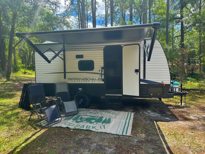 A bright RV rental, with an extended awning & open door, parked at a scenic campsite with chairs and mat.. Keystone RV Springdale 2024