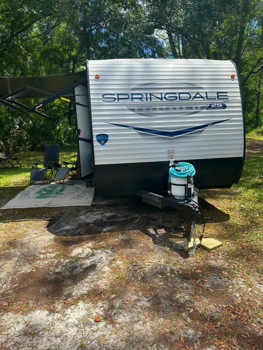 RV rental setup: A white Keystone Springdale travel trailer parked with awning extended, chairs, and mat. Ready for camping!. Keystone RV Springdale 2024