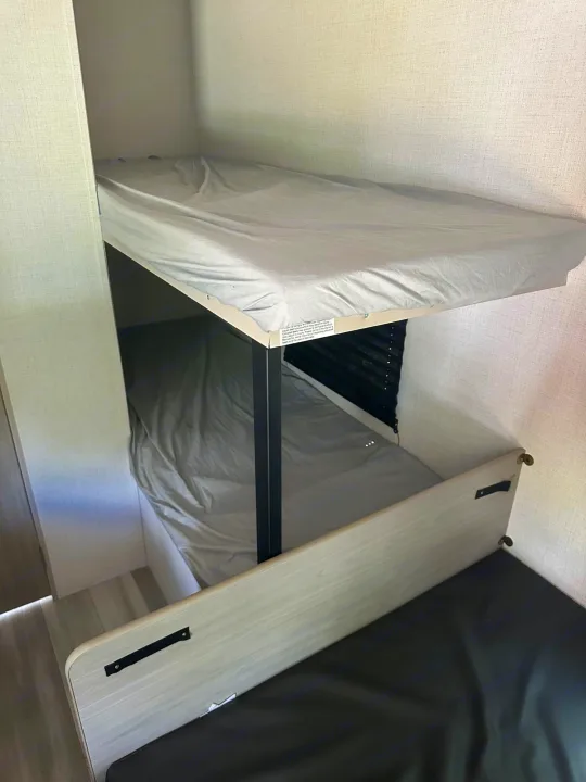 RV rental bunk beds: Two stacked berths with light gray sheets. Below the lower bunk's rail is a dark cushion/mattress.. Keystone RV Springdale 2024
