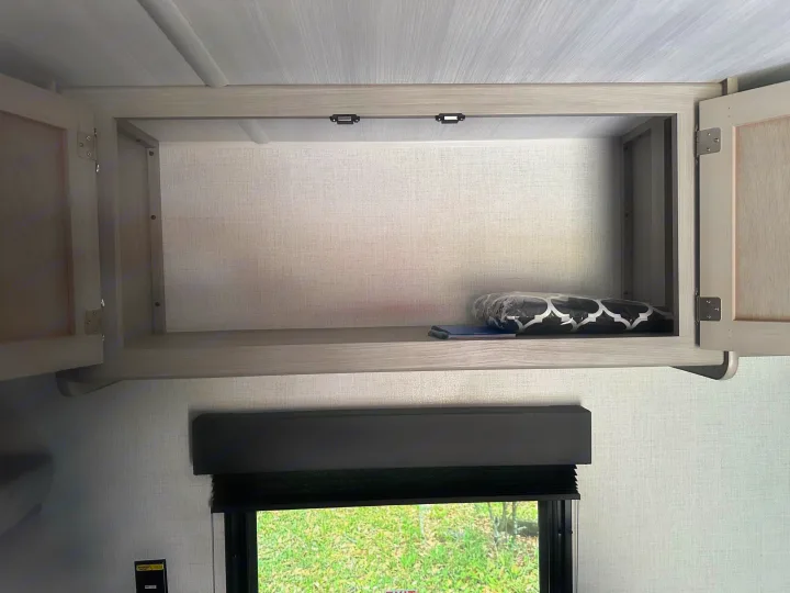 Inside an RV rental: an open overhead cabinet with a shelf, holding a patterned blanket and notebook. A window with a black blind shows green grass below.. Keystone RV Springdale 2024