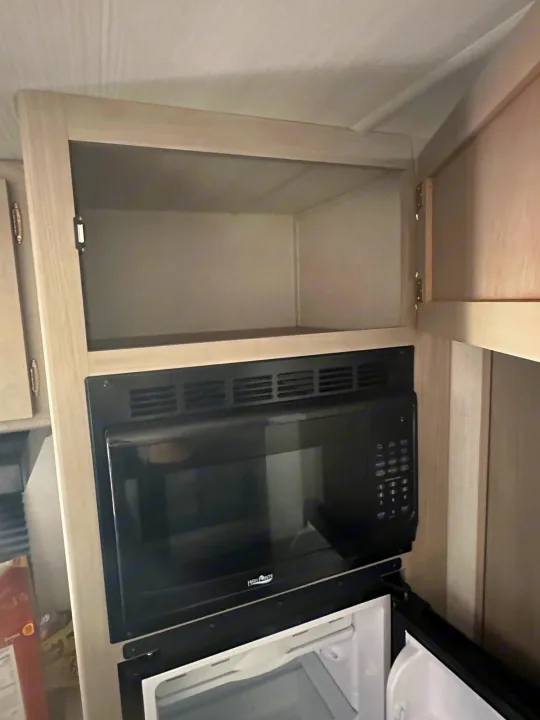 This RV rental's kitchen features an open light-wood cabinet above a black built-in microwave, with a white mini-fridge visible below.. Keystone RV Springdale 2024