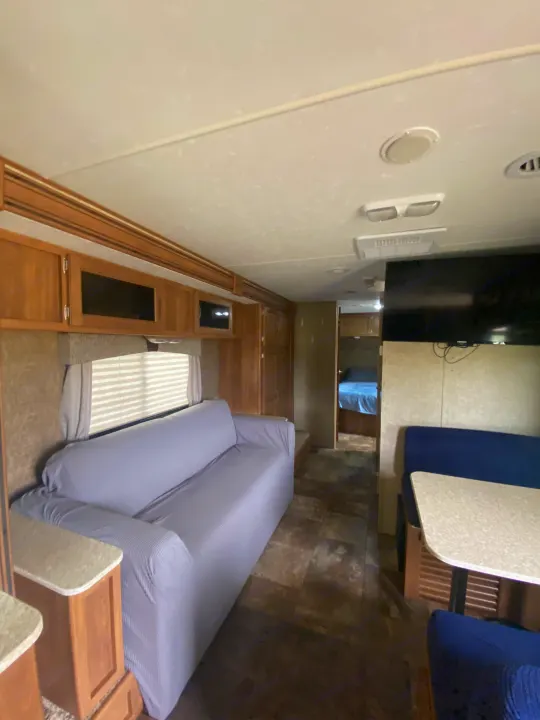 Spacious RV rental interior featuring a grey sofa, blue dining booth, wood cabinets, and a view into the bedroom.. Coachmen Apex 2014