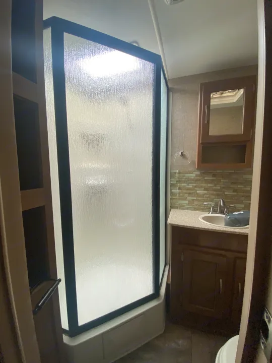 RV rental bathroom: neo-angle shower with frosted glass, facing a vanity with sink, mirrored cabinet, and tiled backsplash.. Coachmen Apex 2014