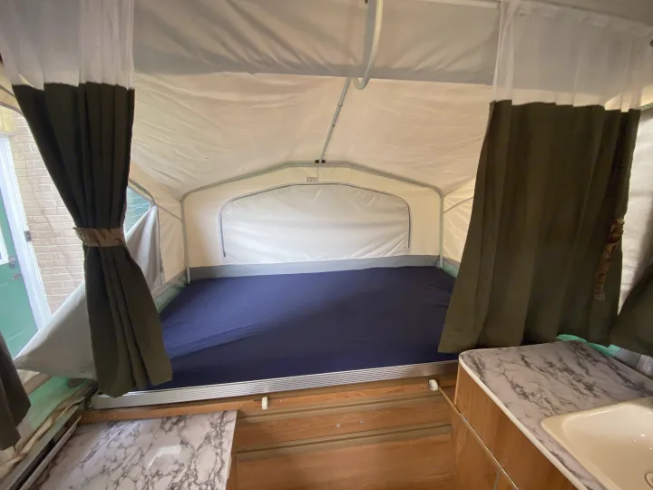 Interior of an RV rental: a pop-up camper's comfortable sleeping area with a blue mattress, green curtains, and partial kitchen.. Palomino Real - Lite 801 2007