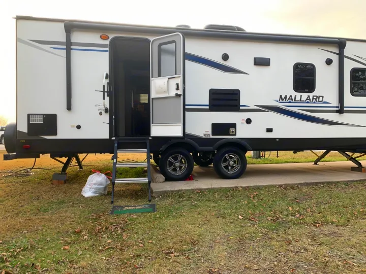 A white Mallard travel trailer, an RV rental, is parked on grass with its door open, steps down, and tandem axles.. Heartland RVs Mallard 2021