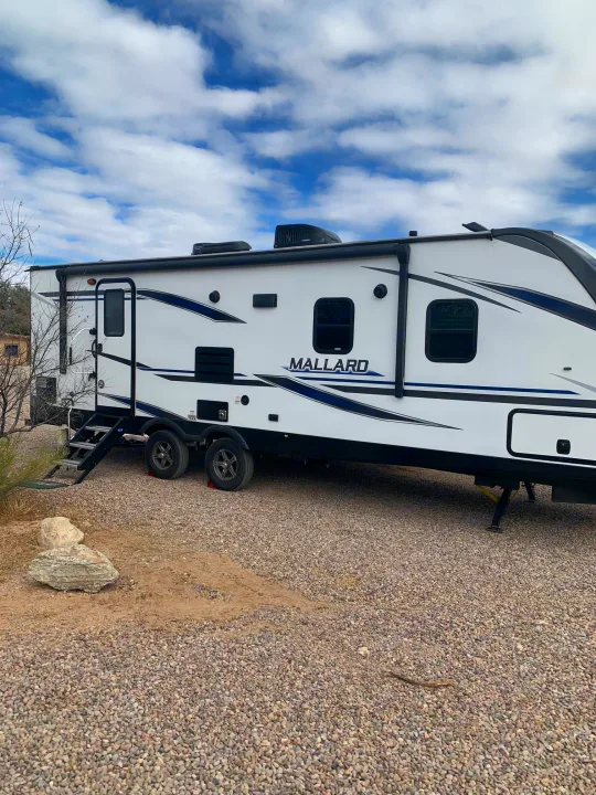 A white Mallard RV rental (travel trailer) is parked on gravel, featuring steps and stripes, under a blue, cloudy sky.. Heartland RVs Mallard 2021