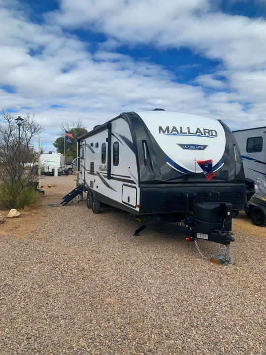 A white Mallard travel trailer RV rental is parked on a gravel site at an RV park, ready for your outdoor adventure.. Heartland RVs Mallard 2021