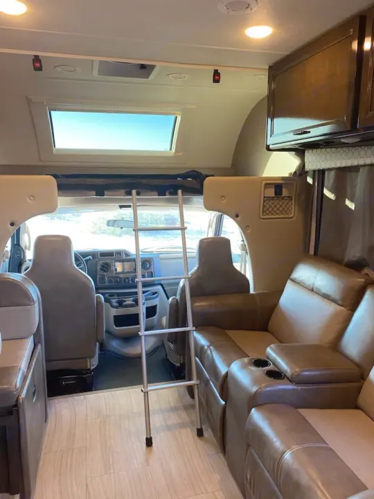 Bright RV rental interior shows the front cab, an overhead bunk bed with a ladder, and comfortable leather recliners.. Thor Quantum 2020