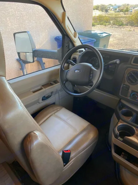 RV rental interior: Driver's seat, Ford steering wheel & dash. Side view shows mirror, bins, and a sunny desert scene.. Thor Quantum 2020