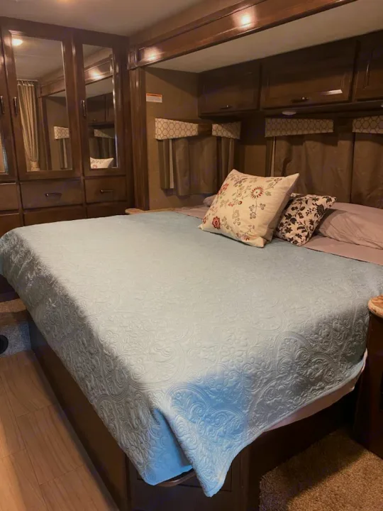 Cozy RV rental bedroom featuring a light blue patterned bedspread, throw pillows, and mirrored storage.. Thor Quantum 2020