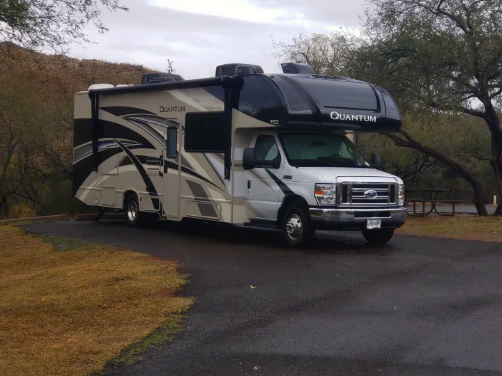 Quantum RV rental, beige and black, parked on asphalt. A picnic table, trees, and a dry hill are visible under a cloudy sky.. Thor Quantum 2020
