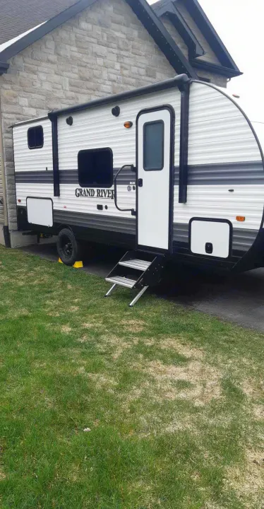 White & black Grand River travel trailer parked by a stone house, showcasing its entrance steps. Perfect for rv rental trips!. Grand River 22MB 2024
