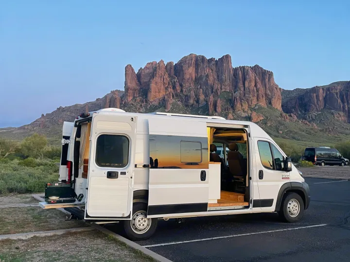 An RV rental, a white campervan with open doors, parked in front of majestic mountains, showcasing its outdoor kitchen.. Dodge Ram ProMaster 2500 2021