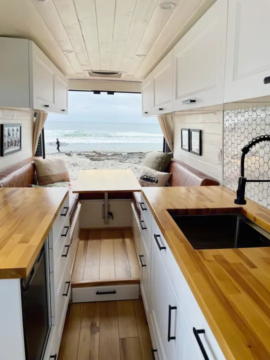 This rv rental shows a bright camper interior with a kitchen, dining area, and an ocean view at a beach with a surfer.. Dodge Ram ProMaster 2500 2021