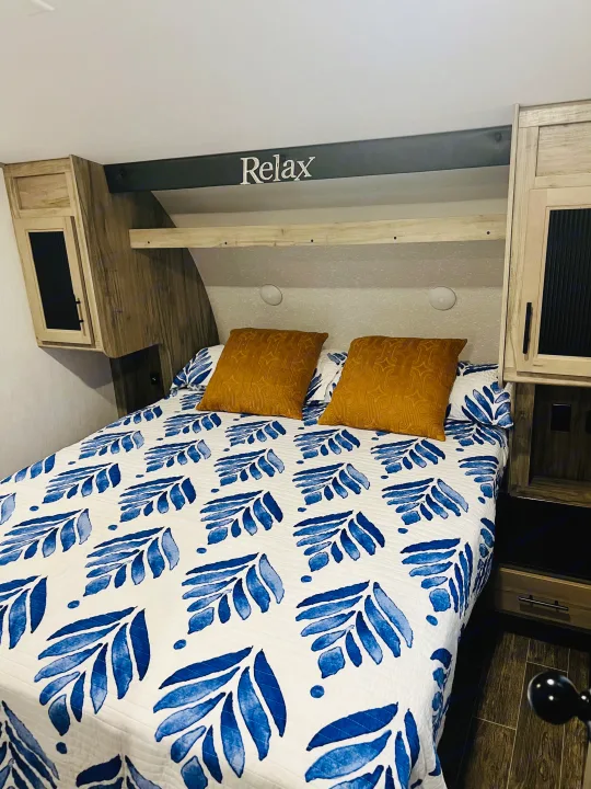 Cozy RV rental bedroom featuring a white and blue leaf-patterned bedspread, golden pillows, and a "Relax" sign.. Forest River Cherokee Arctic Wolf 2021