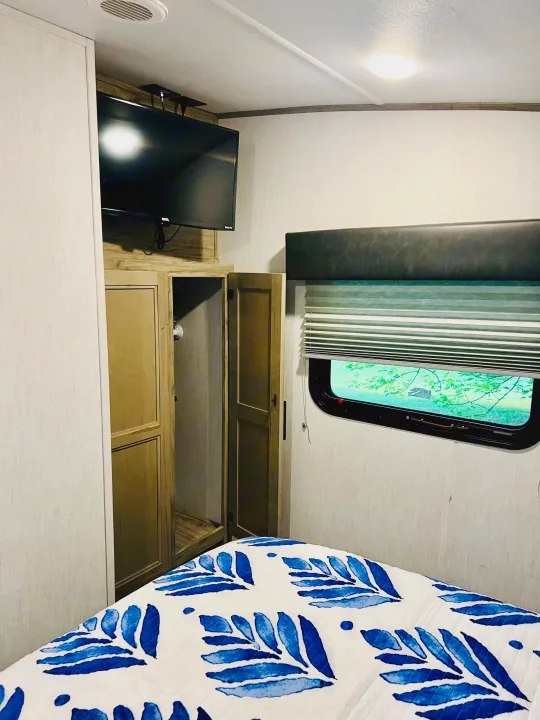 RV rental bedroom with a blue & white patterned bed, wall-mounted TV, open wardrobe, and large window view.. Forest River Cherokee Arctic Wolf 2021