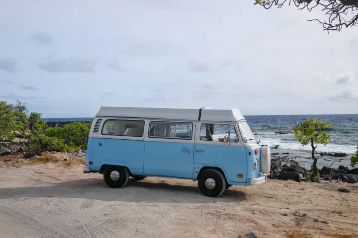 A light blue vintage VW camper van RV rental parked on a sandy beach by the ocean, ready for adventure.. Volkswagen T2 Westfalia 1976