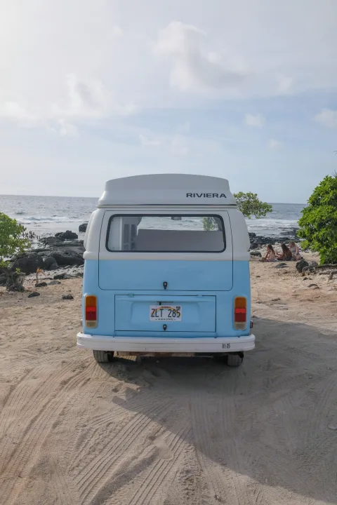 A serene RV rental adventure: a light blue VW camper van parked on a sandy beach beside the ocean, with people relaxing nearby.. Volkswagen T2 Westfalia 1976