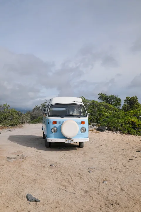 Vintage light blue VW bus rv rental with pop-top, parked on sandy Hawaii track among green bushes under cloudy sky.. Volkswagen T2 Westfalia 1976