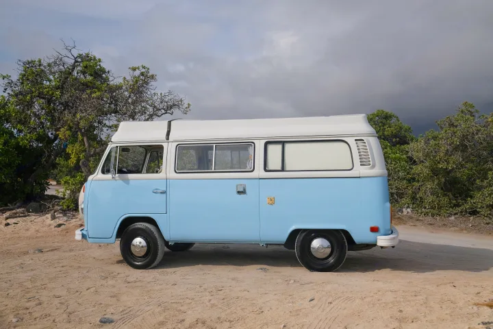 Light blue & white vintage VW camper van on a sandy road by trees under a cloudy sky. Ideal RV rental for your next adventure!. Volkswagen T2 Westfalia 1976