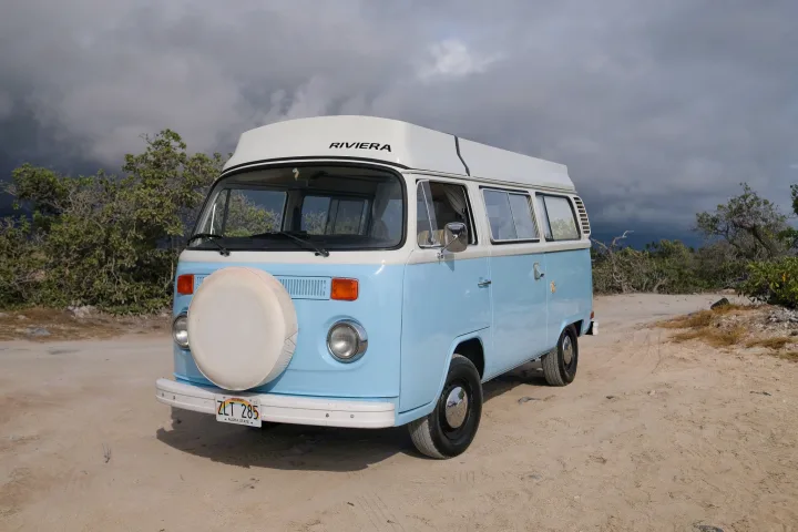 A classic light blue VW camper van with white pop-top and front spare tire, parked on a sandy Hawaii road under dark clouds. Perfect rv rental for adventure.. Volkswagen T2 Westfalia 1976