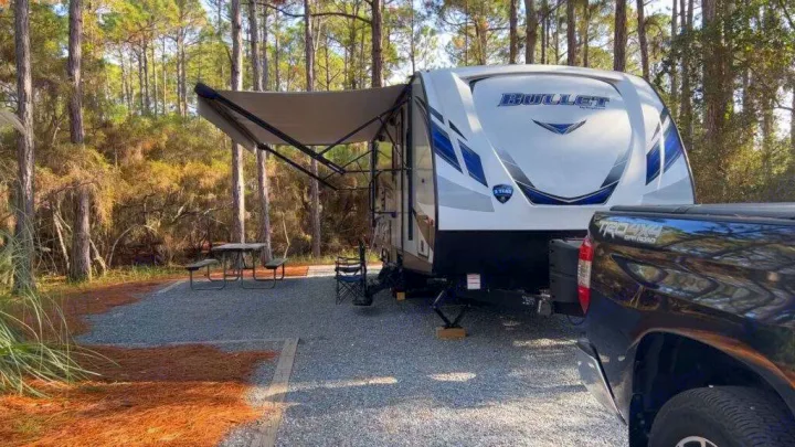 This RV rental is parked at a scenic campsite, with its awning extended beside a truck and picnic table, surrounded by pine trees.. Keystone Bullet Ultra 2018