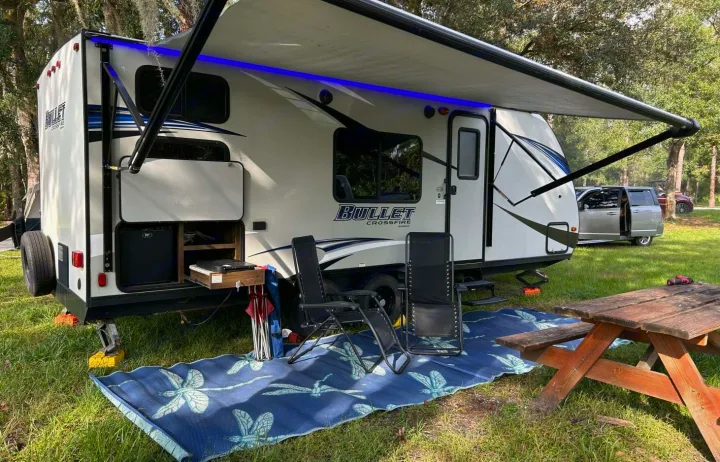 This inviting RV rental campsite features a white travel trailer with an awning, outdoor kitchen, chairs, mat, and picnic table.. Keystone Bullet Ultra 2018