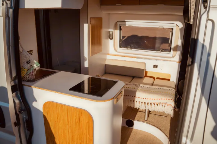 Modern RV rental interior: compact kitchen with cooktop, cozy seating, and a small bathroom, bathed in sunlight.. Noovo Lite 2023