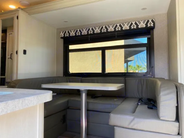 Inside an RV rental: a modern U-shaped dinette with grey cushions, table, and window with a patterned valance, ready for travel.. Entegra Coach Odyssey 2021