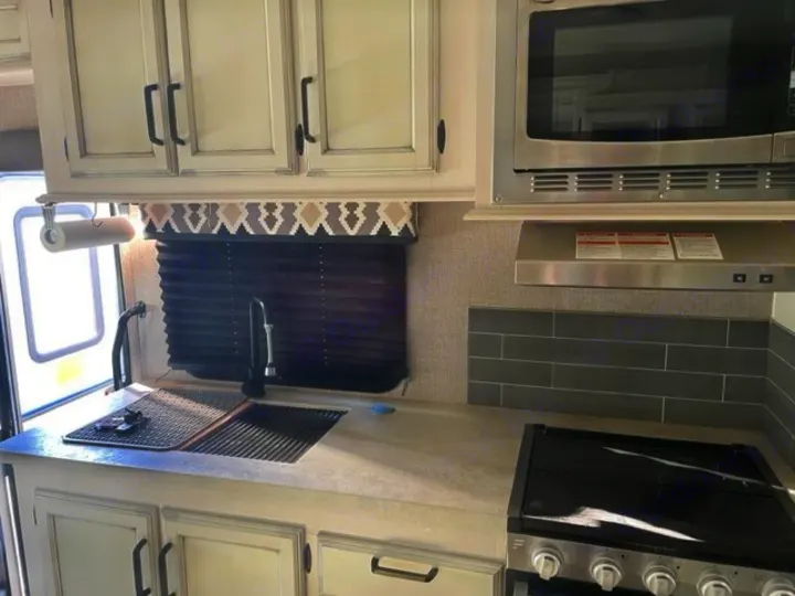 RV rental kitchen: light counters, sink with drying rack, black stove, microwave, white cabinets, and backsplash.. Entegra Coach Odyssey 2021
