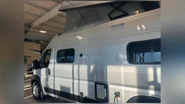 A silver pop-top camper van, perfect for RV rental, is parked indoors, showing its extended roof and side utility ports.. Winnebago Solis 2023