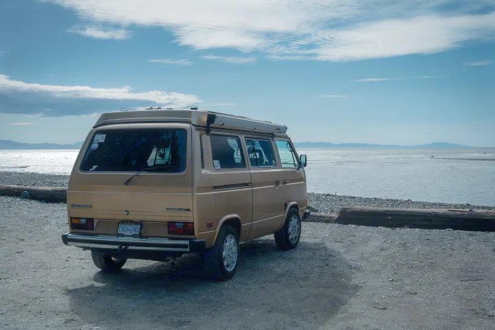 Golden VW Vanagon **RV rental** parked on a gravel beach by the ocean. Distant mountains under a blue sky.. Volkswagen Westfalia 1989