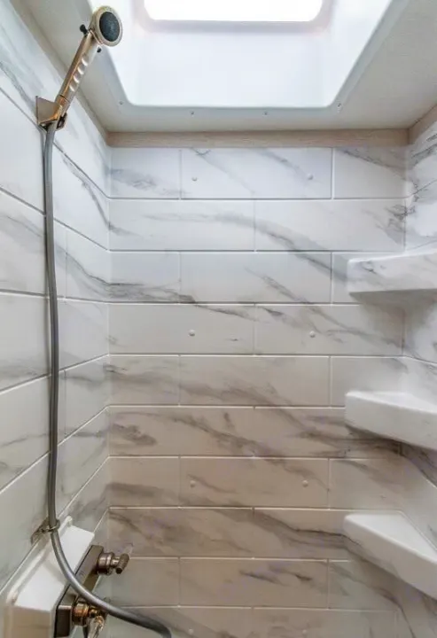 Modern RV rental shower with marble-look tiled walls, handheld showerhead, corner shelves, and a skylight, offering comfort on the go.. Thor Outlaw 2024