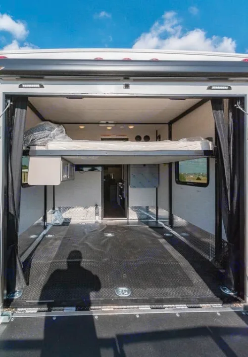 Inside an RV rental toy hauler: A spacious rear garage with a high-mounted bed, durable black floor, and ramp door open to the sky.. Thor Outlaw 2024