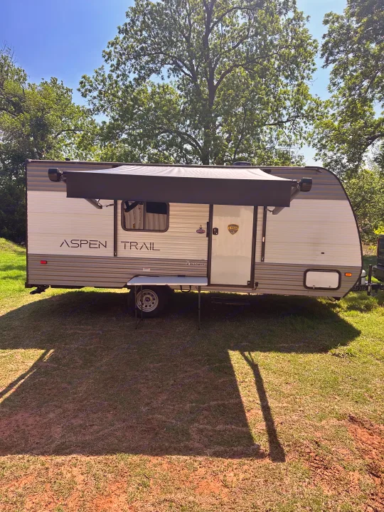 This Aspen Trail RV rental is parked on grass with its awning extended, featuring a small table for outdoor enjoyment under a sunny sky.. Keystone RV Aspen trail 2024