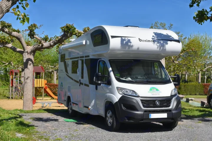 A white Benimar RV rental is parked on a gravel lot next to a tree and children's playground under a clear sky.. Benimar Sport 340 UP 2024