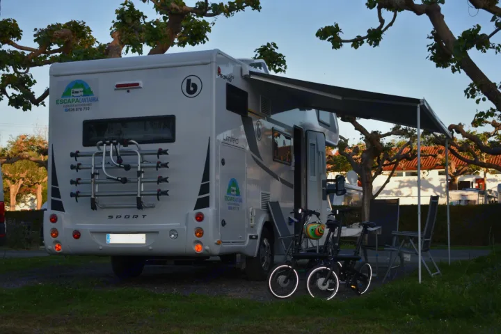 This white motorhome RV rental, branded "ESCAPACANTABRIA", features an extended awning and bicycles at a campsite.. Benimar Sport 340 UP 2024