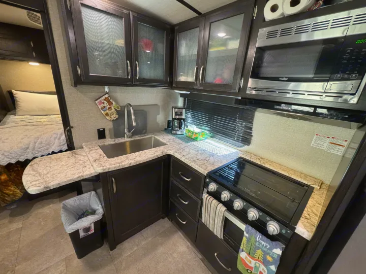 Explore this RV rental's compact kitchen with dark cabinets, a sink, stove, microwave, and adjacent bedroom for comfortable travels.. Grand Design Imagine 2019