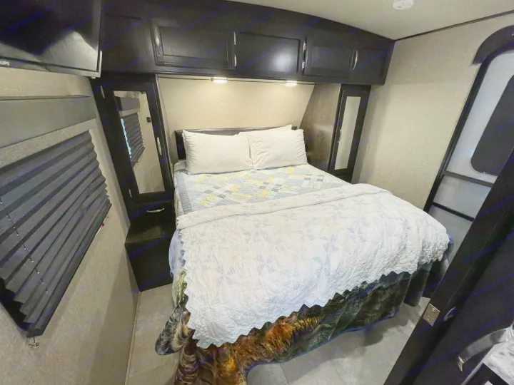 Inside an RV rental, a compact bedroom features a made bed with quilts, overhead cabinets, a TV, and window blinds.. Grand Design Imagine 2019