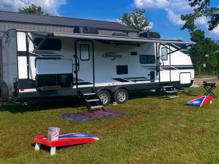 An RV rental parked on a grassy field with awnings extended, steps down, and cornhole boards set up. A grill and building are nearby.. Grand Design Imagine 2019