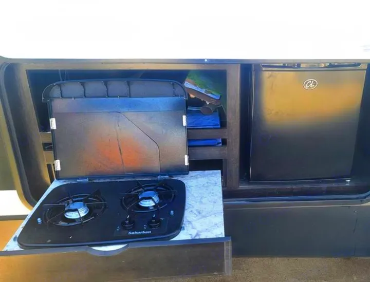 An RV rental's outdoor kitchen, showing a two-burner Suburban gas stove on a pull-out counter and a black mini-fridge.. Grand Design Imagine 2019