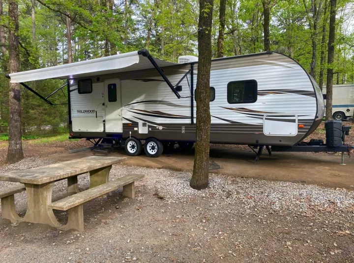 An RV rental travel trailer is parked on a gravel campsite with its awning extended and a concrete picnic table.. Forest River Wildwood 2018
