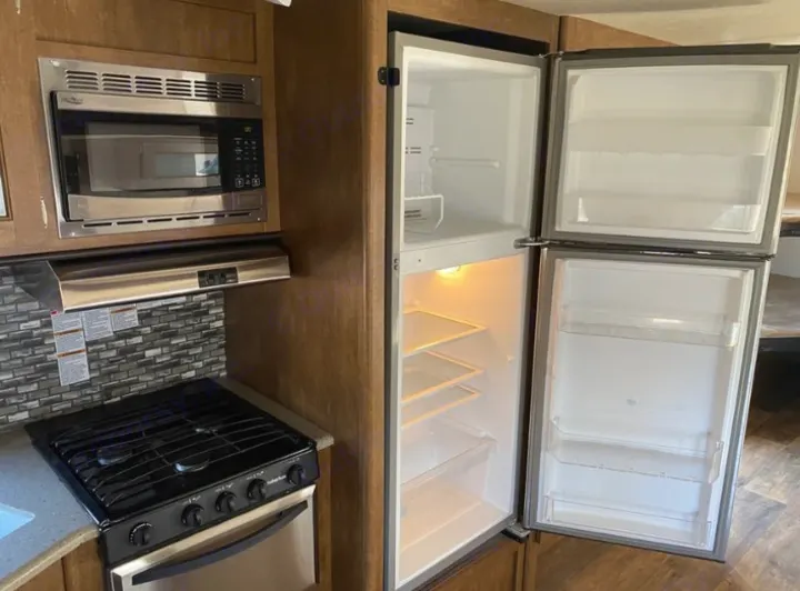 RV rental kitchen with open, empty fridge/freezer, microwave, stove, and oven. Stainless steel appliances, tiled backsplash.. Forest River Wildwood 2018