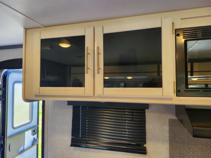 RV rental interior: White overhead glass cabinets with pull handles, an integrated microwave, and dark window blinds.. Crossroads RV Volante 2022