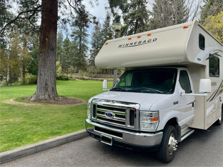 A cream-colored Winnebago rv rental, parked on asphalt next to a park with a large tree and green grass.. Winnebago Minnie Winnie 2017