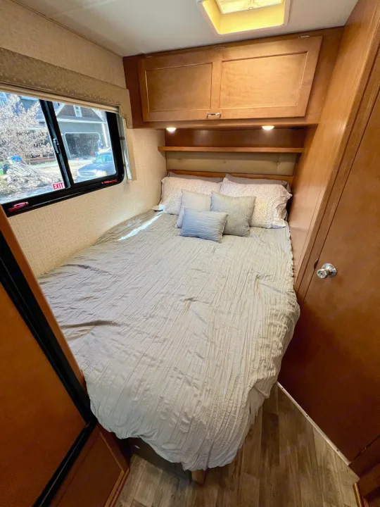 Explore compact living in this RV rental bedroom, featuring a comfy bed, overhead storage, and a scenic window view.. Winnebago Minnie Winnie 2017