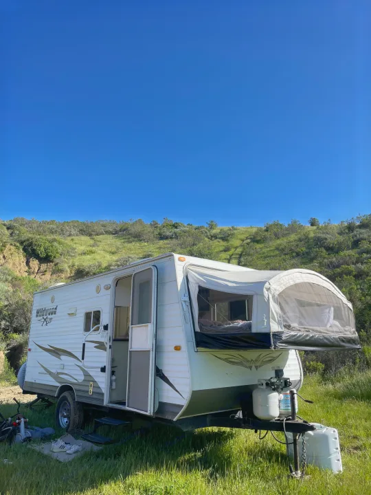 A white rv rental travel trailer with an open door and pop-out bed is set up in a grassy landscape under a clear blue sky.. Forest River Wildwood 2011