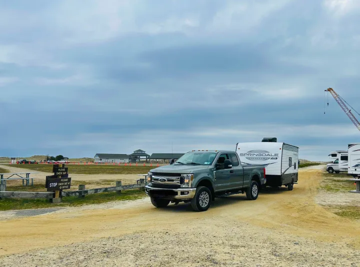 Grey Ford truck towing a Springdale travel trailer, an RV rental, on a dirt road at a campground check-out. Other RVs nearby.. Keystone RV Springdale Mini 1800BH 2024