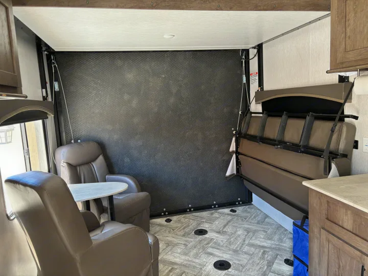 RV rental's adaptable toy hauler interior, featuring recliners, table, and a fold-up bed against a textured wall.. Forest River Stealth 2022