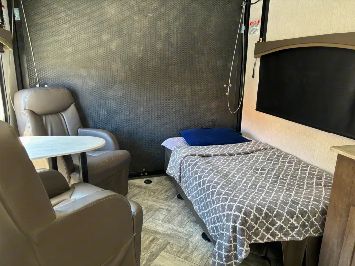 Inside an RV rental: two gray recliners, a small table, and a bed with a patterned blanket.. Forest River Stealth 2022